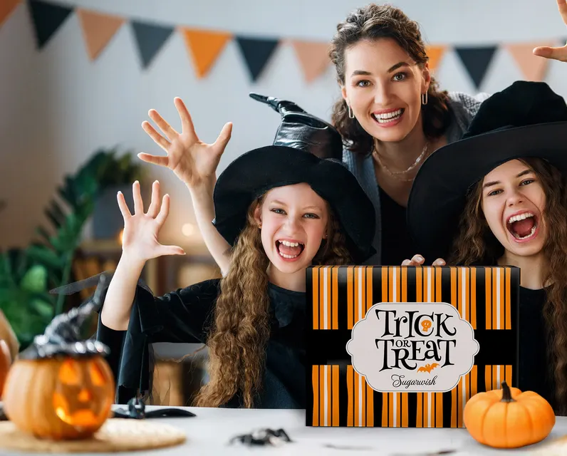Mom and two daughters in witch hats laughing while holding a Trick-or-Treat Sugarwish gift box, surrounded by Halloween decorations and pumpkins