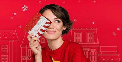 Woman subtly smiling and waving a Sugarwish holiday box in front of holiday shops